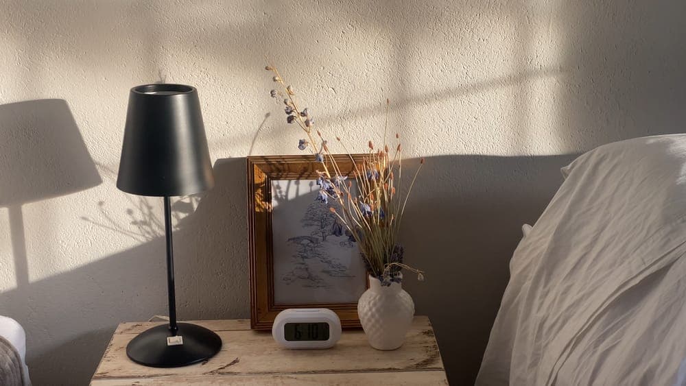 A sunlit wooden nightstand with a lamp, a clock and dried flowers beside a pillow