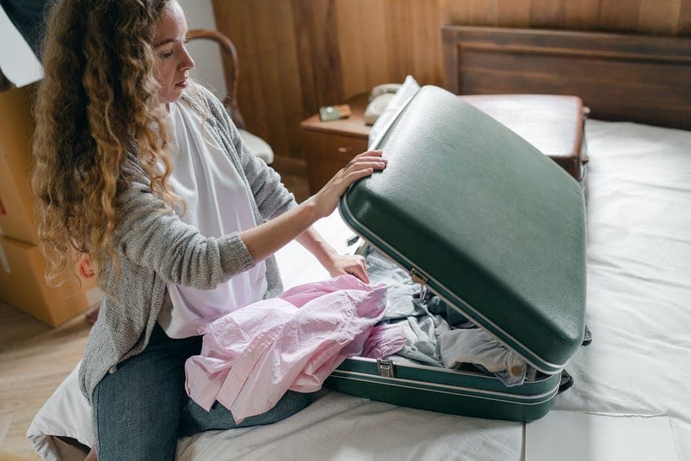 A woman packing clothes into an open green suitcase on a bed