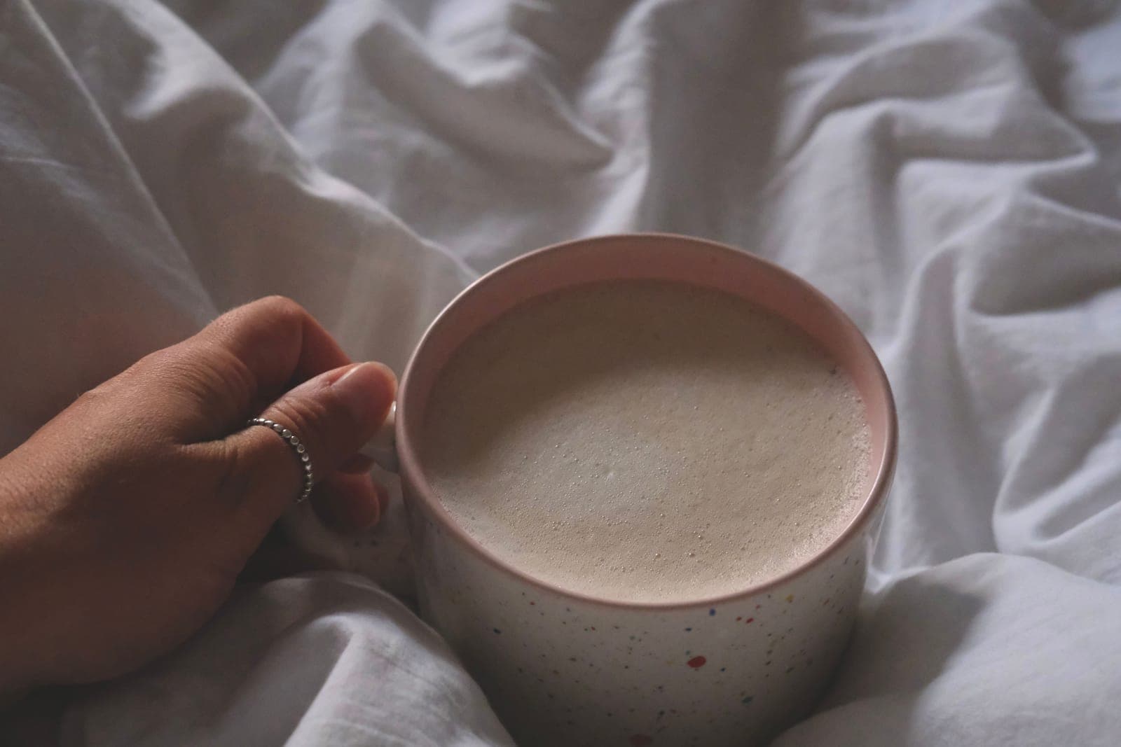 A hand with a thin silver ring holds a mug of frothy coffee on rumpled white bedsheets